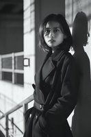 Young Asian woman in elegant trench coat standing by concrete wall with dramatic shadows in black and white portrait for fashion and lifestyle content.