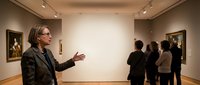 Professional museum guide presenting artwork to diverse group of visitors in modern gallery space with warm lighting and classical paintings on walls.
