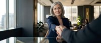 Professional mature businesswoman in dark suit shaking hands across conference table in modern office with city view windows during corporate meeting.
