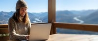 Young woman in knit sweater and beanie working on laptop at mountain cabin with scenic alpine valley view through large windows during winter vacation.