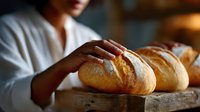 Fresh crusty bread loaves on rustic wooden board with baker's hands checking quality in warm natural lighting, artisanal bakery atmosphere.