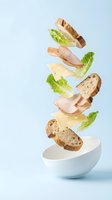 Flying sandwich ingredients falling into white bowl with ham cheese lettuce and artisan bread slices on light blue background for food advertising.