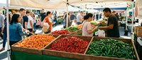 Busy outdoor farmers market with colorful fresh vegetables, diverse shoppers browsing stalls with peppers, cucumbers, and tomatoes on a sunny day.