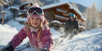 Young girl in pink ski jacket and goggles smiling while sliding down snowy slope with wooden chalet and another skier in background. Winter family vacation.