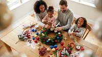 Multiracial family with two girls crafting a Christmas wreath at a wooden table with colorful ornaments and festive decorations.