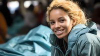 Young African American woman with blonde curly hair smiling warmly while wearing teal winter coat outdoors. Natural daylight creates soft bokeh background effect.