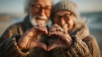 Senior couple making heart shape with hands on beach at sunset showing love and togetherness in golden hour light for romantic lifestyle concepts.