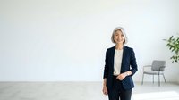 Mature woman with white hair and glasses wearing a black blazer over a white shirt, standing confidently in a modern office space with minimalist decor and plants.