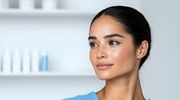Young Hispanic woman with glowing natural makeup and sleek dark hair in blue top against white spa background, showcasing fresh dewy skin and confident smile.
