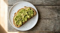 Mashed avocado toast on artisan bread served on white plate with black pepper seasoning on rustic wooden table background for healthy breakfast menu.