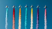 Colorful airplanes flying in formation against blue sky leaving vibrant smoke trails in turquoise, red, orange, yellow, purple and pink colors creating artistic pattern.