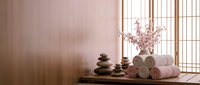 Zen spa setting with stacked stones, rolled towels and cherry blossoms on wooden surface near traditional window screen for wellness and relaxation.