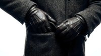 Businessman in dark charcoal wool coat wearing black leather gloves with quilted pattern, hands positioned in coat pockets, cropped torso view against white background for corporate and fashion use.