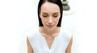 Young woman with long dark hair and fair complexion wearing white sleeveless top, looking downward with calm expression in studio setting for healthcare, wellness, or professional use.