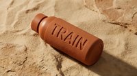 Terracotta water bottle with embossed train text resting on sandy beach surface, suitable for travel, lifestyle, and product design projects.