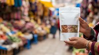 Vendor holding transparent pouch with dried spices at outdoor market stall, displaying product for sale with blurred marketplace background.