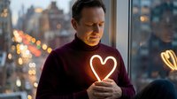 Middle-aged Caucasian man in burgundy sweater holding glowing heart symbol by window with city lights bokeh background at evening for love concepts.