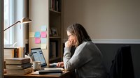 Young woman working from home office with laptop computer and books on desk near window. Remote work productivity and study concept.