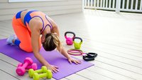 Young athletic woman in colorful sportswear stretching on purple yoga mat with kettlebells and dumbbells on wooden deck for home fitness workout routine.