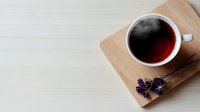 Minimalist composition with steaming cup of dark tea on wooden board with delicate lavender sprig against light wooden surface. Cozy morning atmosphere.