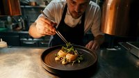 Chef with beard and light skin tone plating gourmet dish with tweezers in modern kitchen, garnishing food with microgreens for fine dining restaurant presentation.