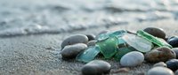 Sea glass and smooth pebbles scattered on sandy beach shore with gentle ocean waves in background creating peaceful coastal scene for relaxation themes.