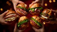 Hands holding gourmet sandwiches on crusty bread with meat, cheese and fresh greens arranged in circle against dark background with bokeh lights.