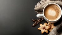 Aromatic cappuccino with heart-shaped foam served with star-shaped cookies and scattered coffee beans on rustic burlap fabric against dark background.