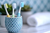 Colorful toothbrushes in decorative blue ceramic holder against soft bathroom background with white towel and green plant, morning hygiene concept.