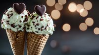 Mint chocolate chip ice cream cones with heart-shaped chocolate toppers against blurred bokeh lights background, romantic dessert setting for Valentine's Day celebration.