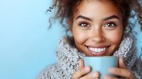 Young mixed-race woman with curly hair smiling while holding light blue mug, wearing cozy knit scarf against soft blue background for lifestyle content.
