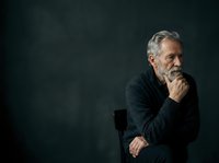 Mature man with gray hair and beard, wearing dark sweater, sitting thoughtfully with hand to chin against dark background, conveying contemplation and wisdom for business, leadership, and professional concepts.