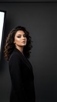 Professional Middle Eastern businesswoman in black suit with curly hair and makeup posing confidently in modern office studio for corporate branding.