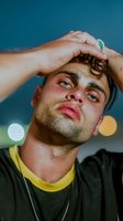 Young Caucasian male with dark hair and facial hair wearing yellow and black shirt, hand on forehead in stressed or tired pose against blue background with bokeh lights.
