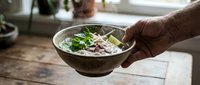 Man's hand holding ceramic bowl with fresh Asian noodle soup garnished with herbs and vegetables, ready to serve for lunch or dinner meal preparation.