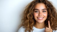 Young woman with curly brown hair and bright smile wearing white top against light background, touching face gently. Natural beauty portrait.