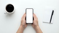 Hands holding smartphone with blank white screen on clean white desk workspace with coffee cup and notepad for mockup presentations.