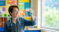 Young Asian female scientist in denim shirt examining test tube with blue liquid in bright laboratory with colorful educational posters on wall.
