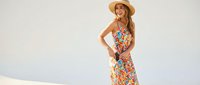 Young woman with long reddish-blonde hair and fair complexion wearing colorful floral print sundress and straw hat, standing confidently against white background for fashion and lifestyle content.