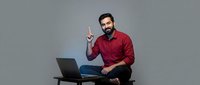 Smiling bearded Middle Eastern businessman in red shirt pointing up while sitting with laptop on desk against gray background for presentations.