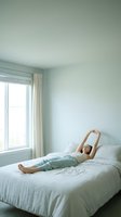 Woman relaxing on bed in bright, minimalist bedroom with natural light from window, stretching arms overhead in comfortable home interior setting.