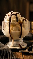 Vanilla ice cream scoop in glass bowl with chocolate syrup drizzle, served on wooden table with spoon for dessert and food photography.