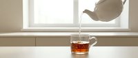 Hot tea being poured from white ceramic teapot into glass cup on minimalist white table near bright window, cozy morning ritual.