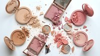 Professional makeup collection featuring pink and nude cosmetics including blush, eyeshadow, and powder compacts scattered on white background.