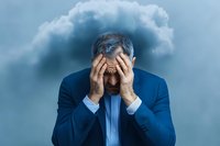 Stressed mature businessman in blue suit covering face with hands against stormy cloud background, representing anxiety, burnout and mental pressure.