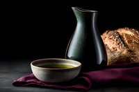 Ceramic bowl with olive oil and black pottery jug on dark background with artisan bread and burgundy linen napkin, moody food photography composition.