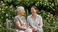 Senior woman with gray hair and adult daughter with brown hair, both Caucasian, sitting together on wooden bench in blooming garden, holding hands, smiling and laughing during quality time outdoors.