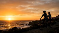 Silhouetted cyclists riding mountain bikes on coastal cliffs at golden hour sunset, with ocean waves and dramatic sky, suitable for sports and outdoor lifestyle content.