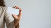 Woman with medium skin tone holding blank white business card in hand wearing beige sweater, ideal for corporate branding and professional networking presentations.