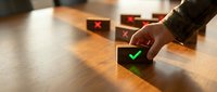 Hand placing wooden cube with green checkmark among blocks with red X marks on desk, representing quality control and selection process in business decisions.